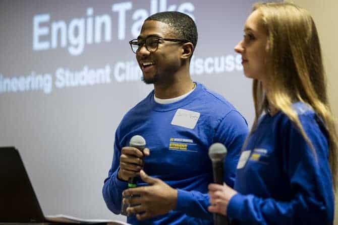 Two students holding microphones stand in front of a projected slide reading "Engin Talks."