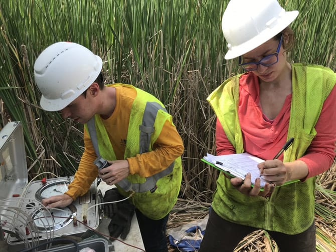 Two students wearing hard hats and visibility vests monitor testing equipment and take notes on data.