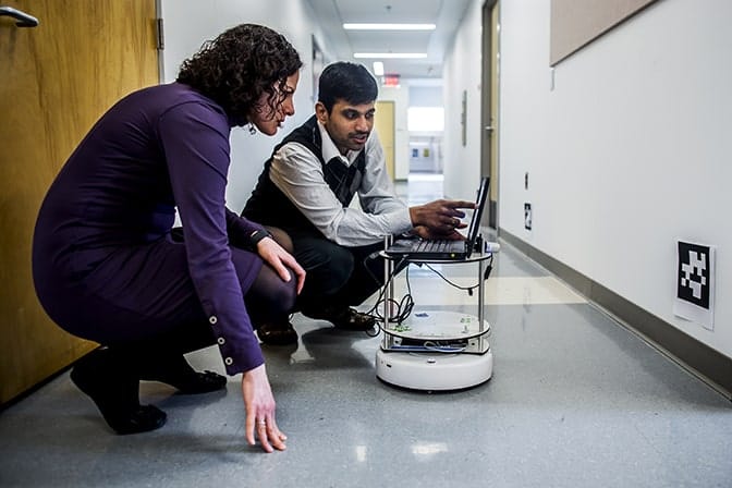 Two people crouch in front of a laptop, with one of them pointing to the screen