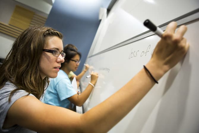 Three people write on a whiteboard at the front of a lecture hall