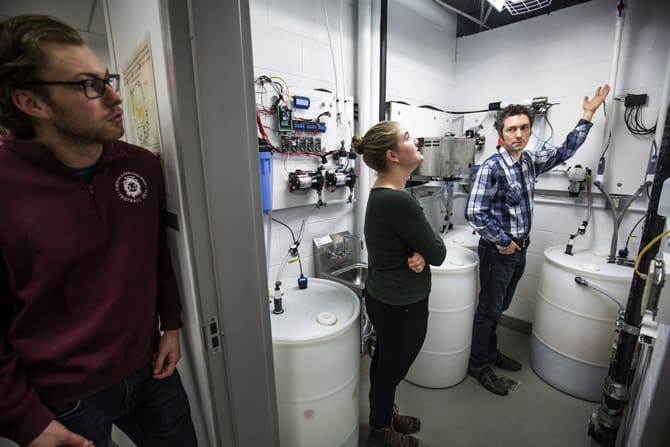 Two people stand in a urine treatment room, with a third person looking in from the door One person gestures towards one of the various pipes running along the walls