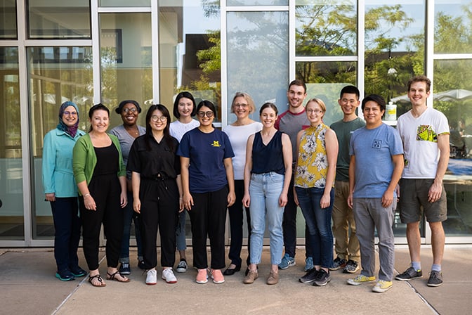 Group of 13 people pose in front of a glass building