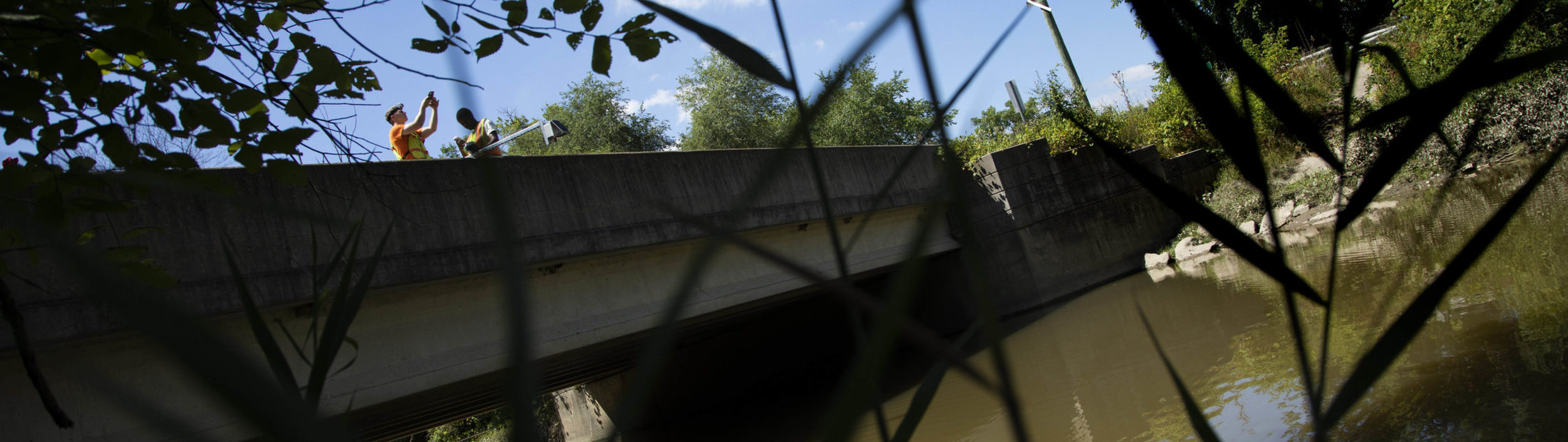 Two students in reflective vests stand on a car bridge next to a metal rod holding a sensor pointed at the water below.