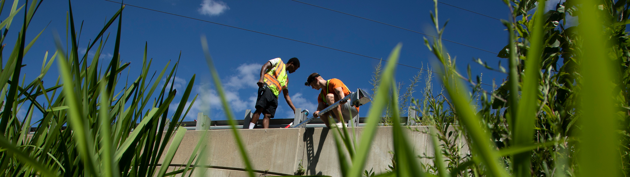 Two researchers stand on top of a bridge adjusting equipment to measure water levels below