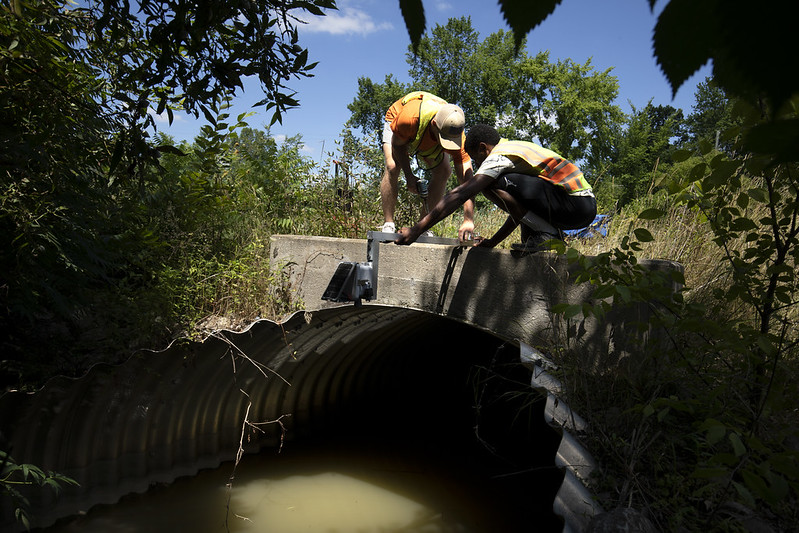 Two researchers stand on top of a bridge adjusting equipment to measure water levels below